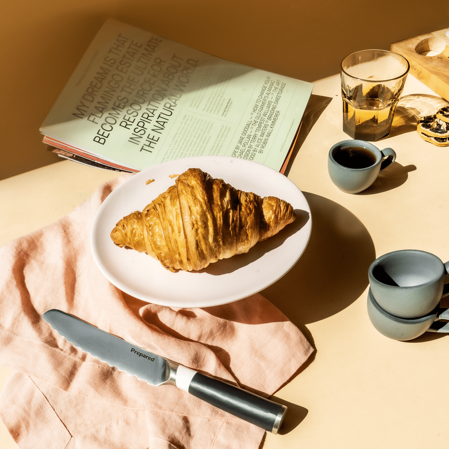 The bread knife resting on a pink table napkin with a delicious croissant and espresso cup of coffee ready with a magazine on the side